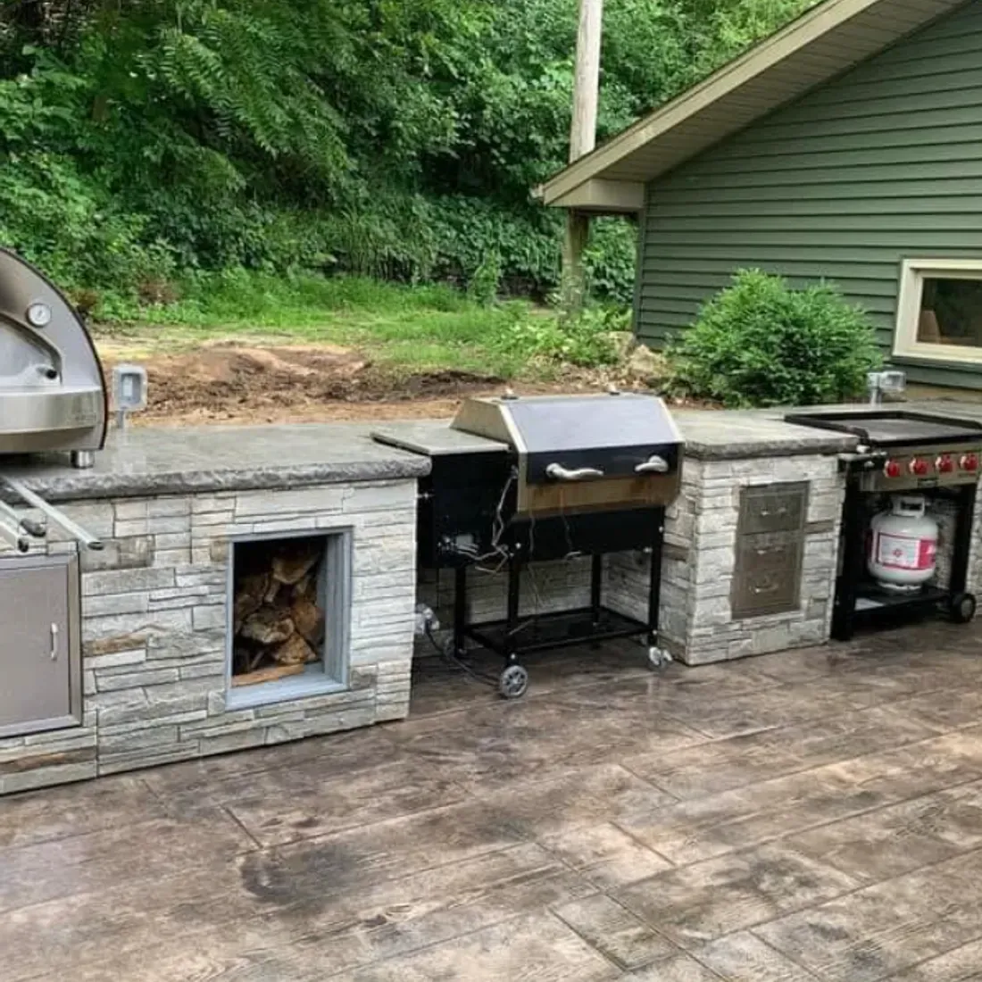 Outdoor kitchen with grill, wood storage, and countertops on a stamped concrete patio.