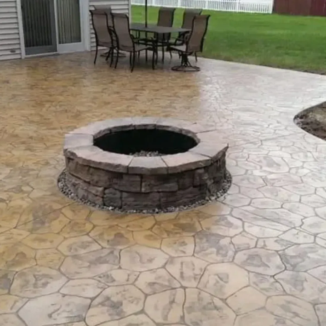 Stamped concrete patio with a stone fire pit, dining table, chairs, and green lawn.