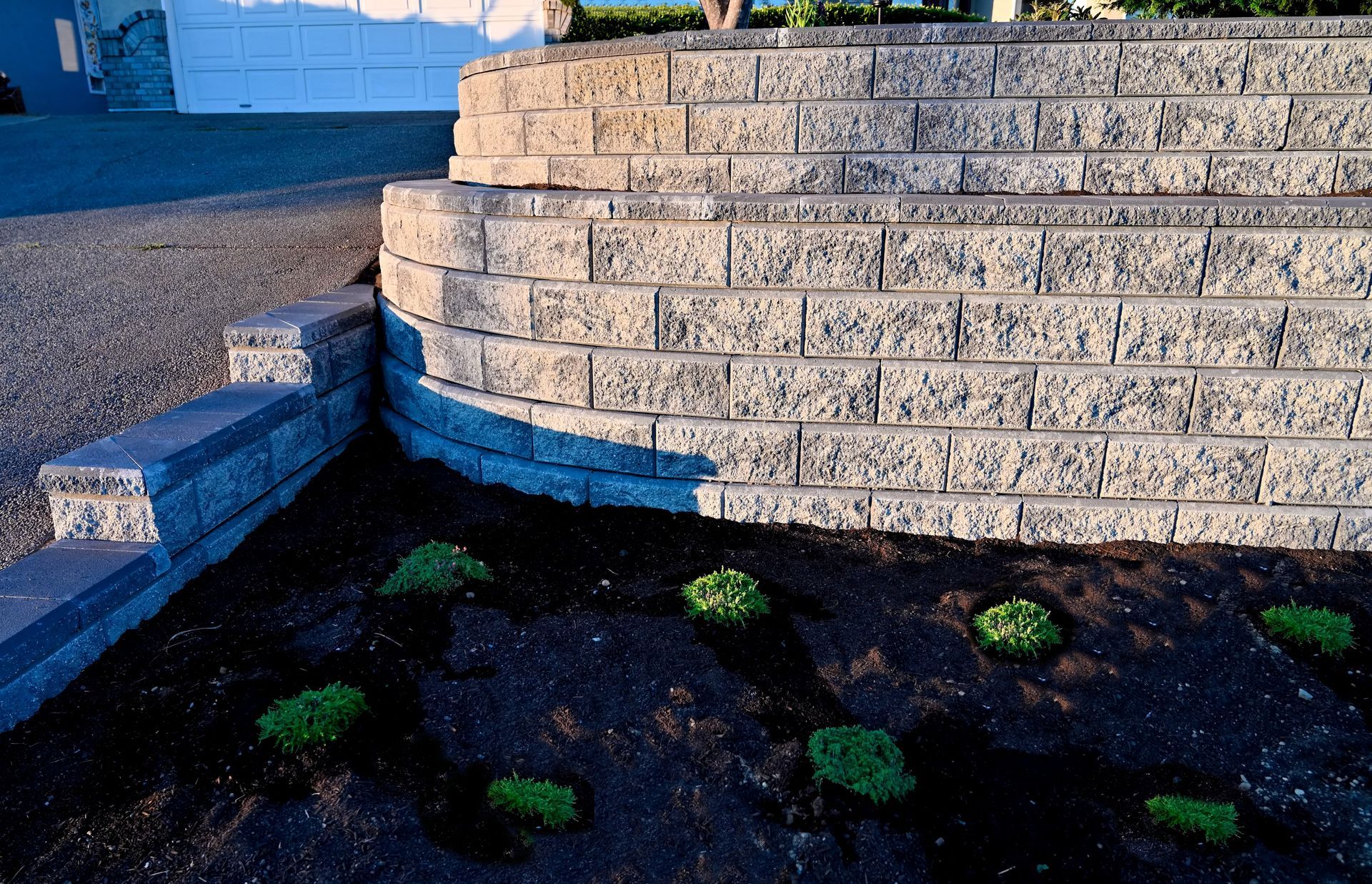 Retaining wall made of textured gray bricks, curved, bordering a garden bed with green plants.