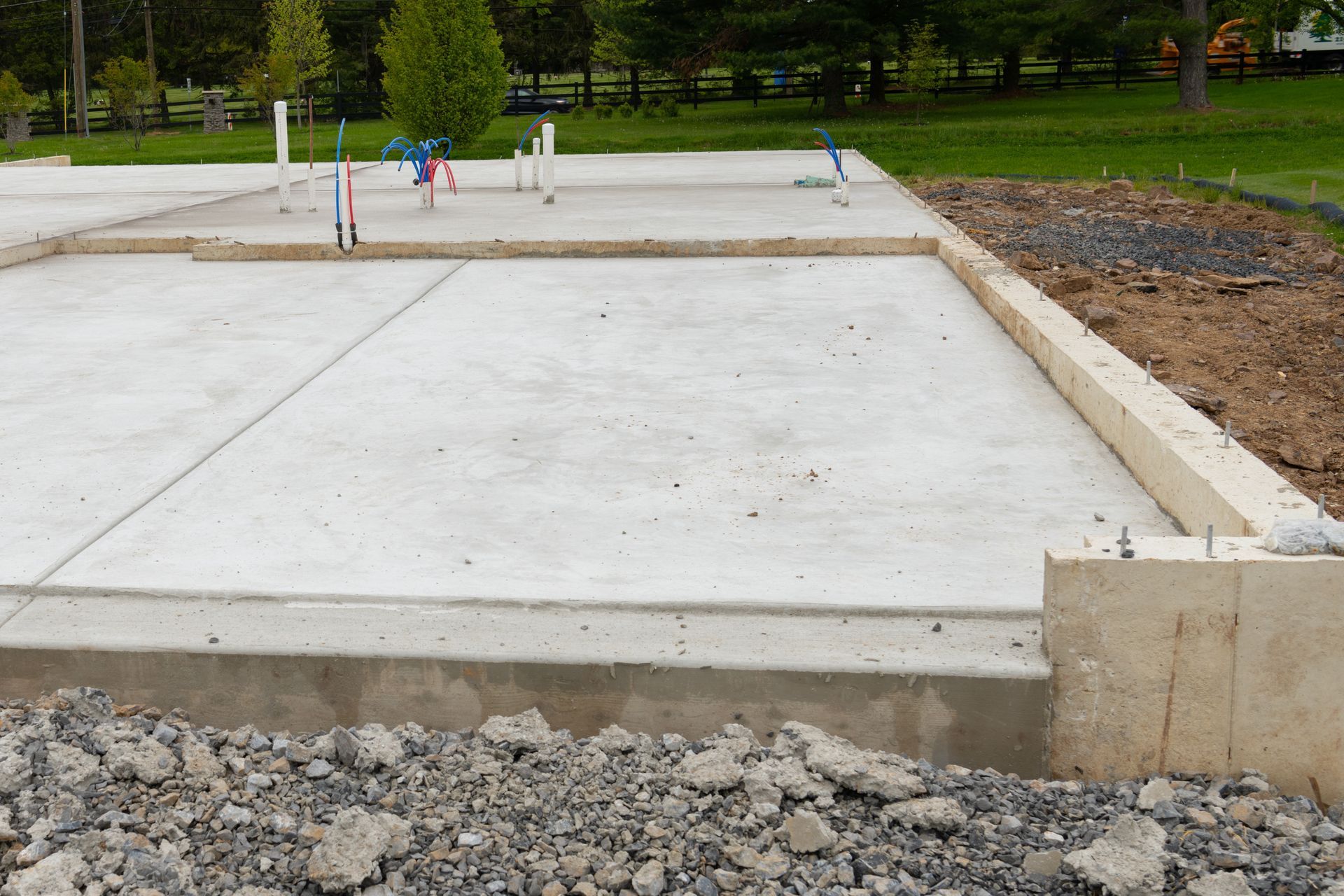 Concrete foundation for a building under construction; plumbing pipes visible, gravel pile in foreground.