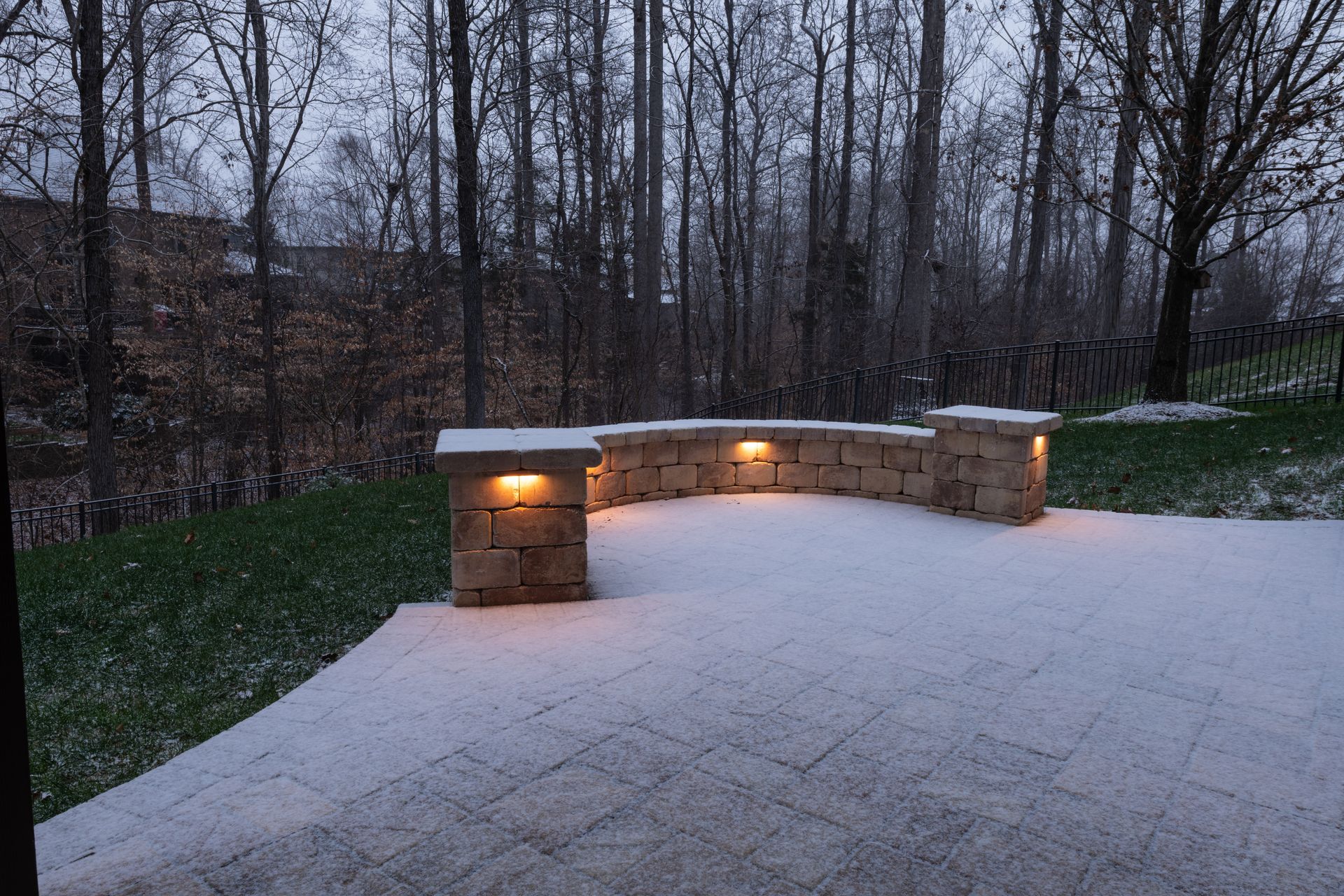 Snow-covered patio with lit stone wall and backdrop of trees and a snowy lawn.