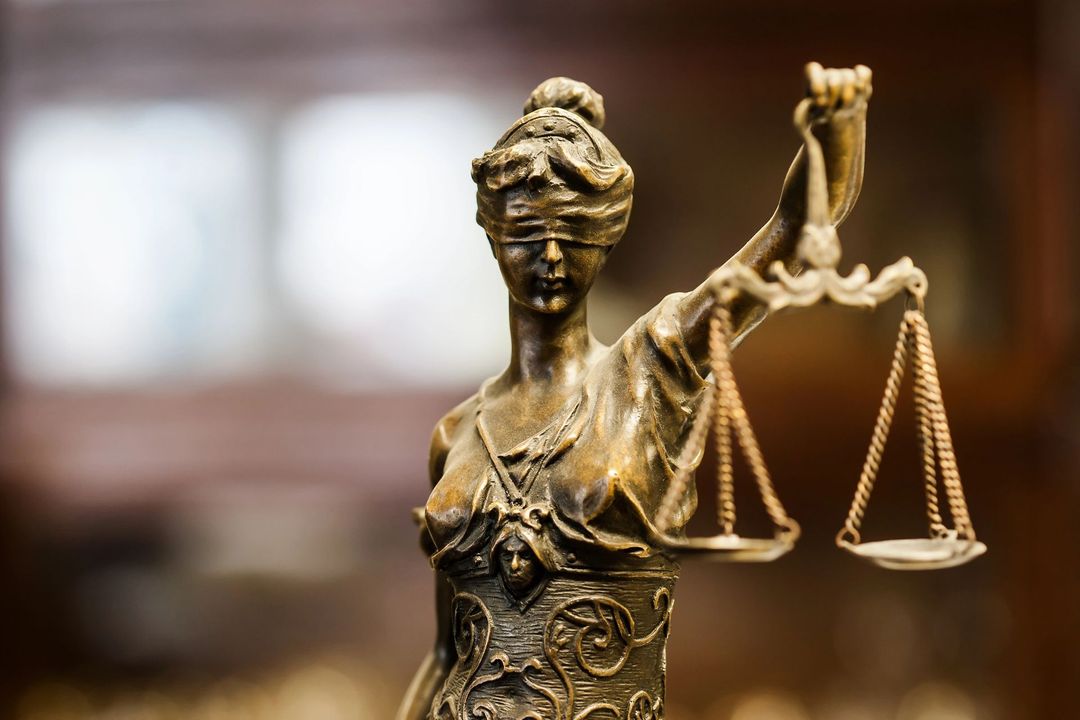 Statua in bronzo della Dea della Giustizia che regge una bilancia, con uno sfondo sfocato di un'aula di tribunale.