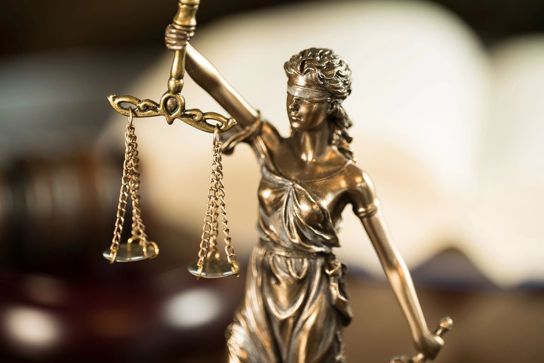 Statua in bronzo della Dea della Giustizia che regge una bilancia, con un libro di legge aperto sfocato sullo sfondo.