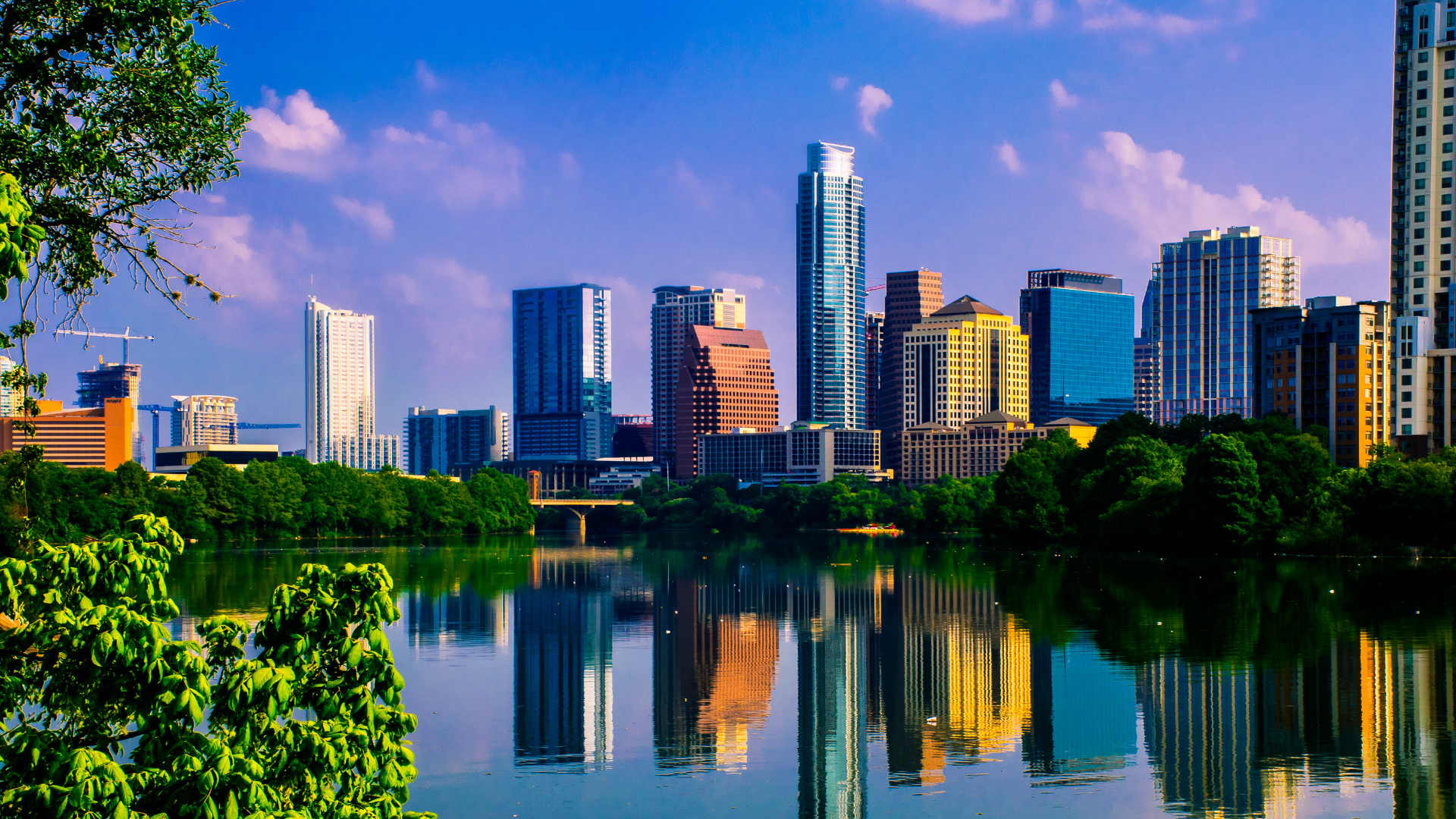 A city skyline is reflected in a lake with trees in the foreground.