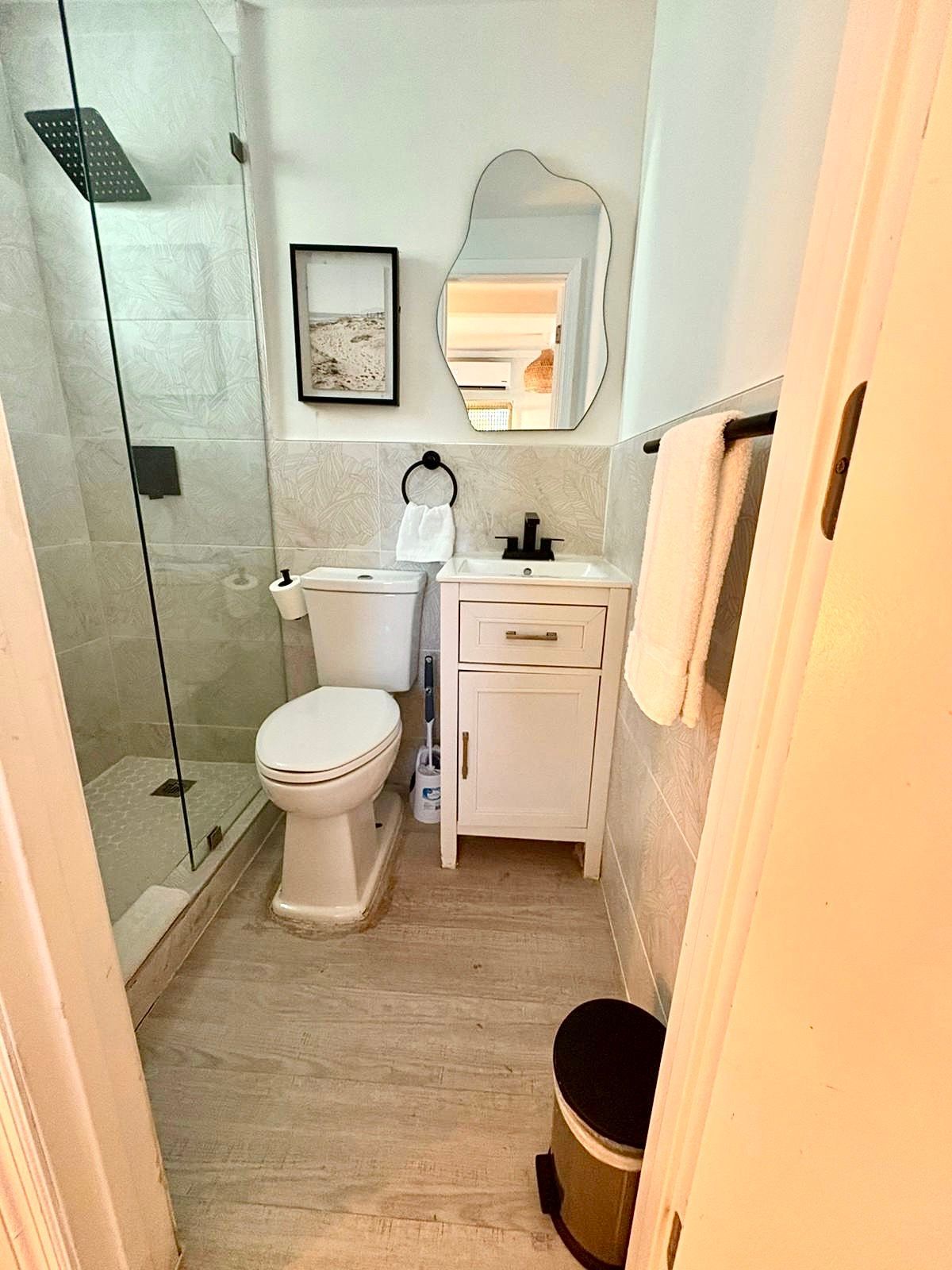 Small, white bathroom with shower, toilet, sink, and mirror. Beige-tiled walls and floors, with black accents.