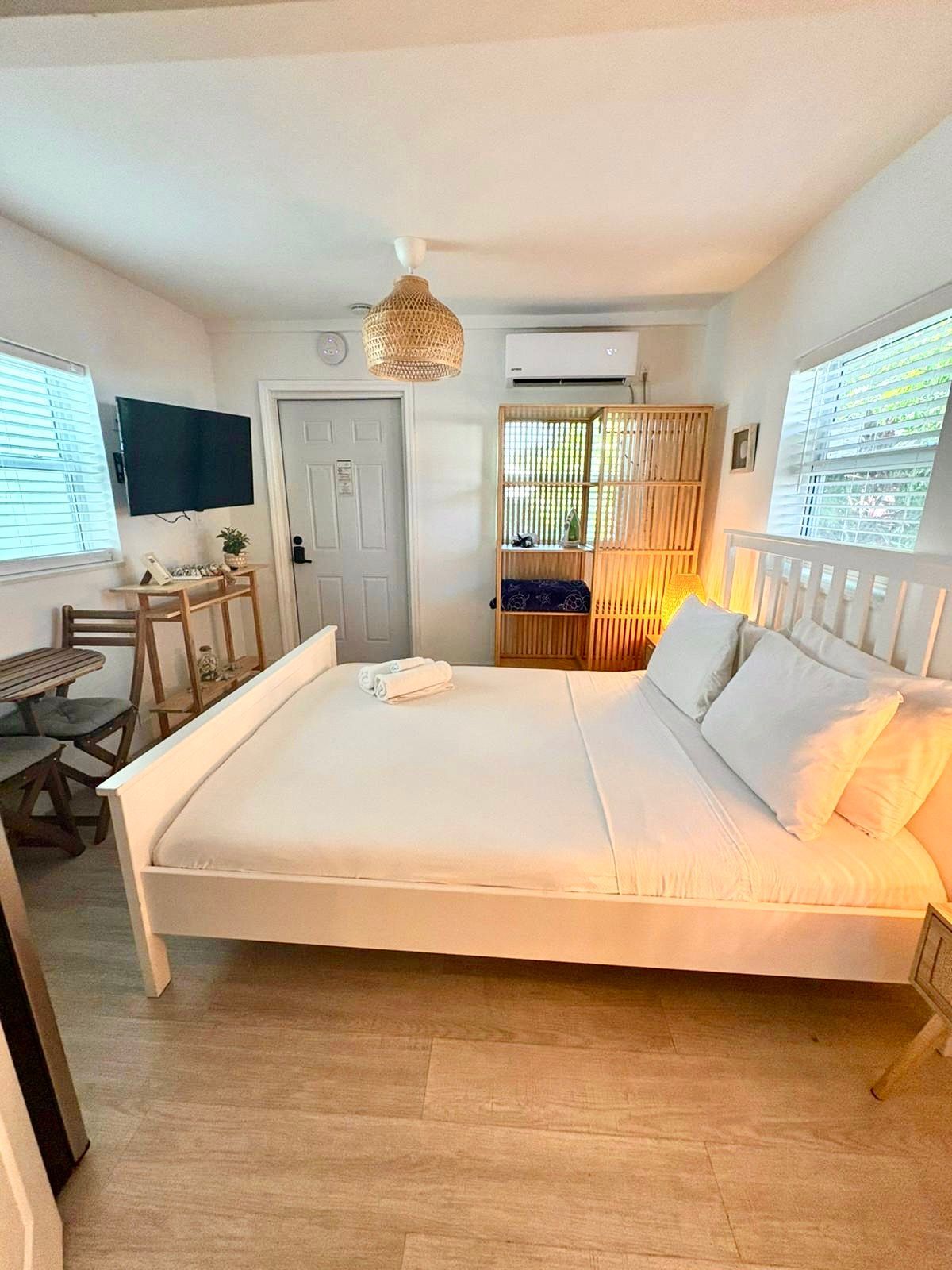 A white bedroom with a bed, TV, small table, and windows.