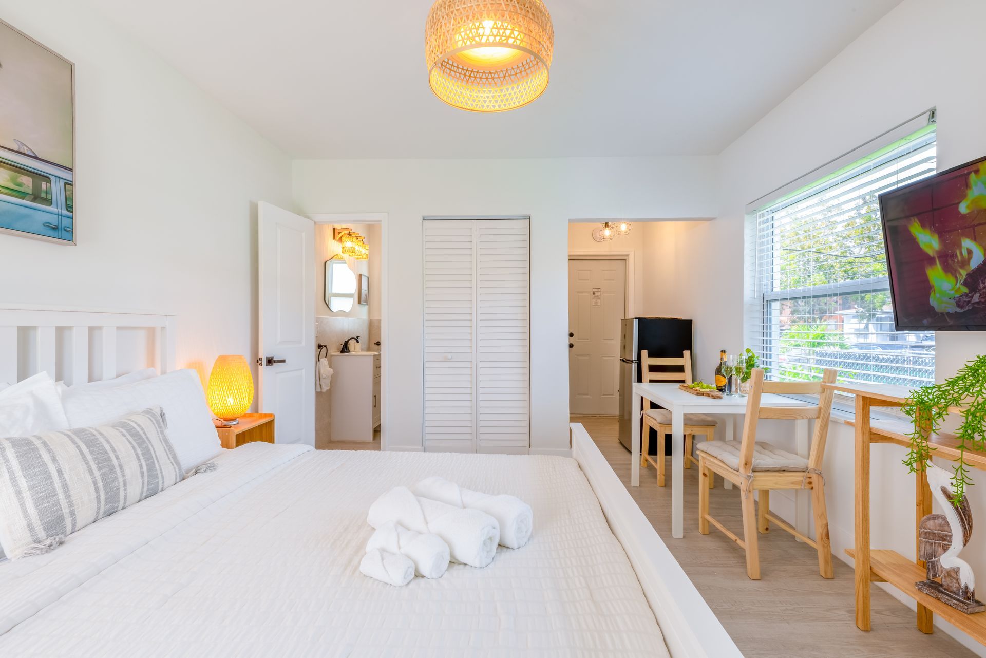 White bedroom with bed, table, bathroom, and television.