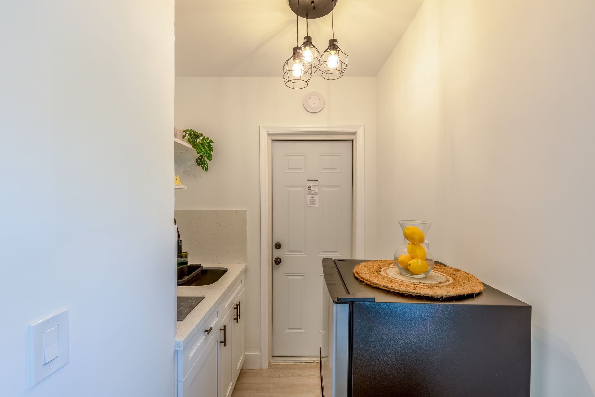 Small kitchen with white cabinets, a mini-fridge, and a door at the back. A glass of juice sits on the fridge.