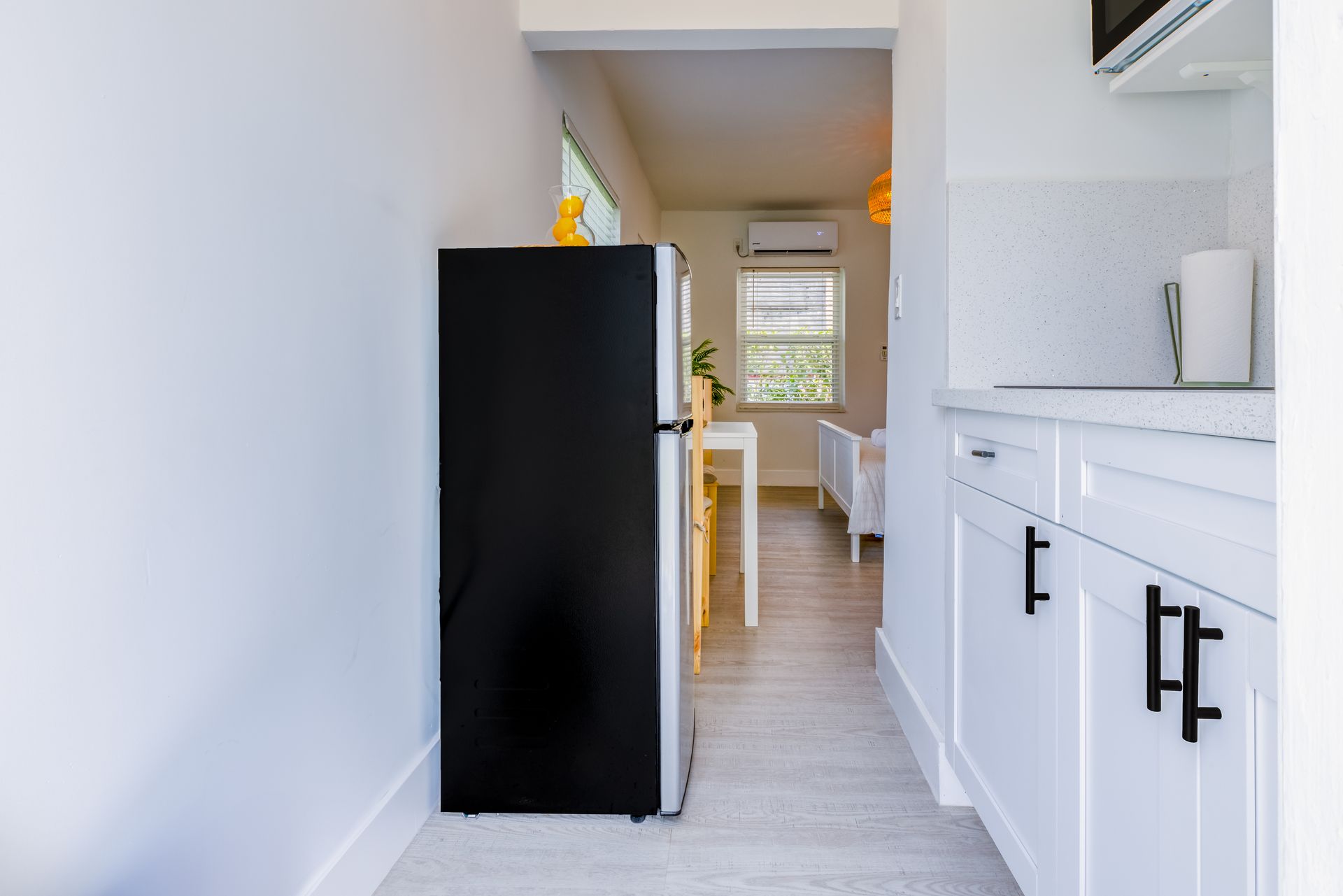 A narrow kitchen hallway with a black refrigerator, white cabinets, and an open doorway to the living area.