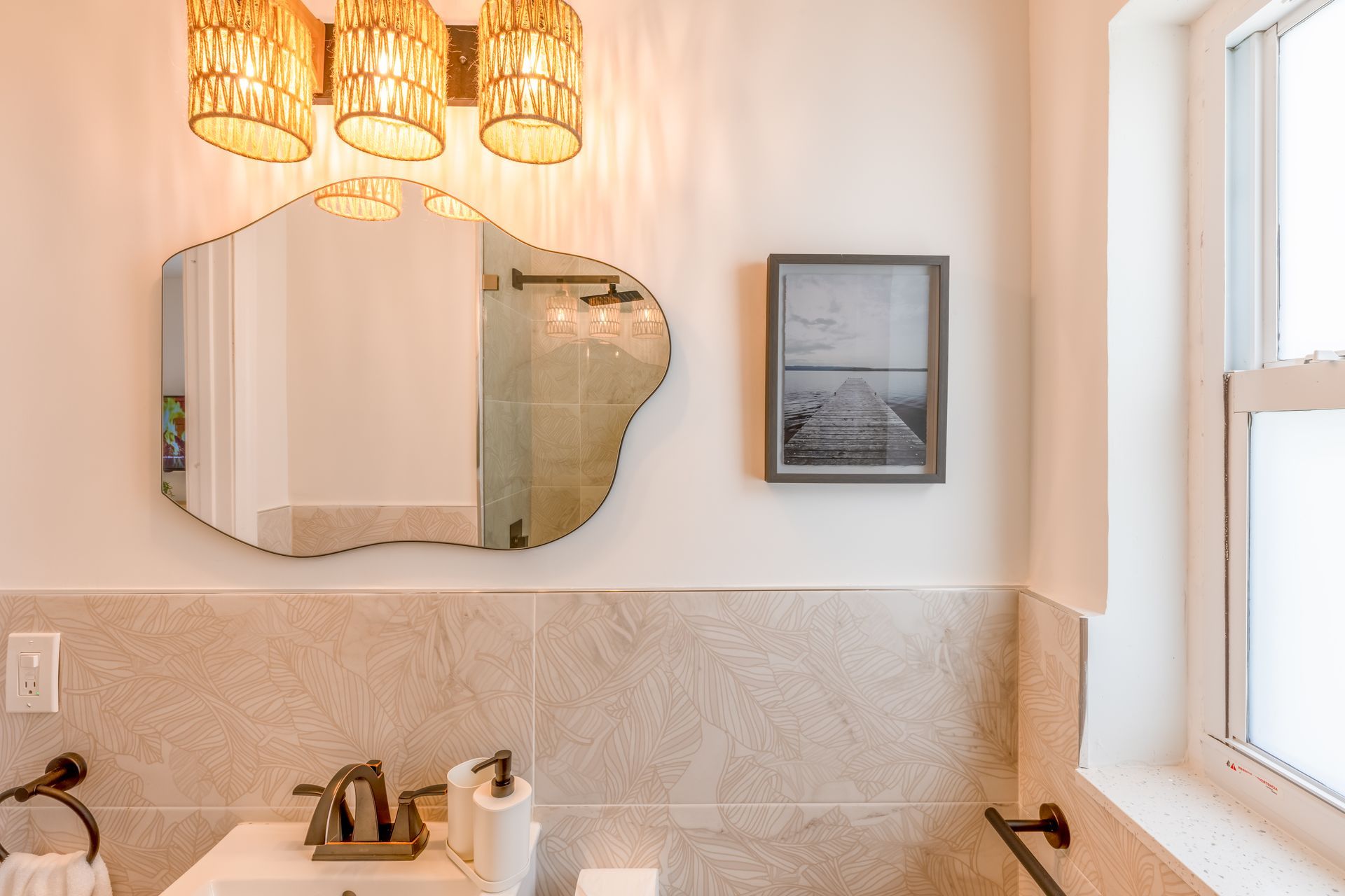 Bathroom with wavy-edged mirror, framed art, and woven light fixture. Beige tile, white walls, and window to the right.