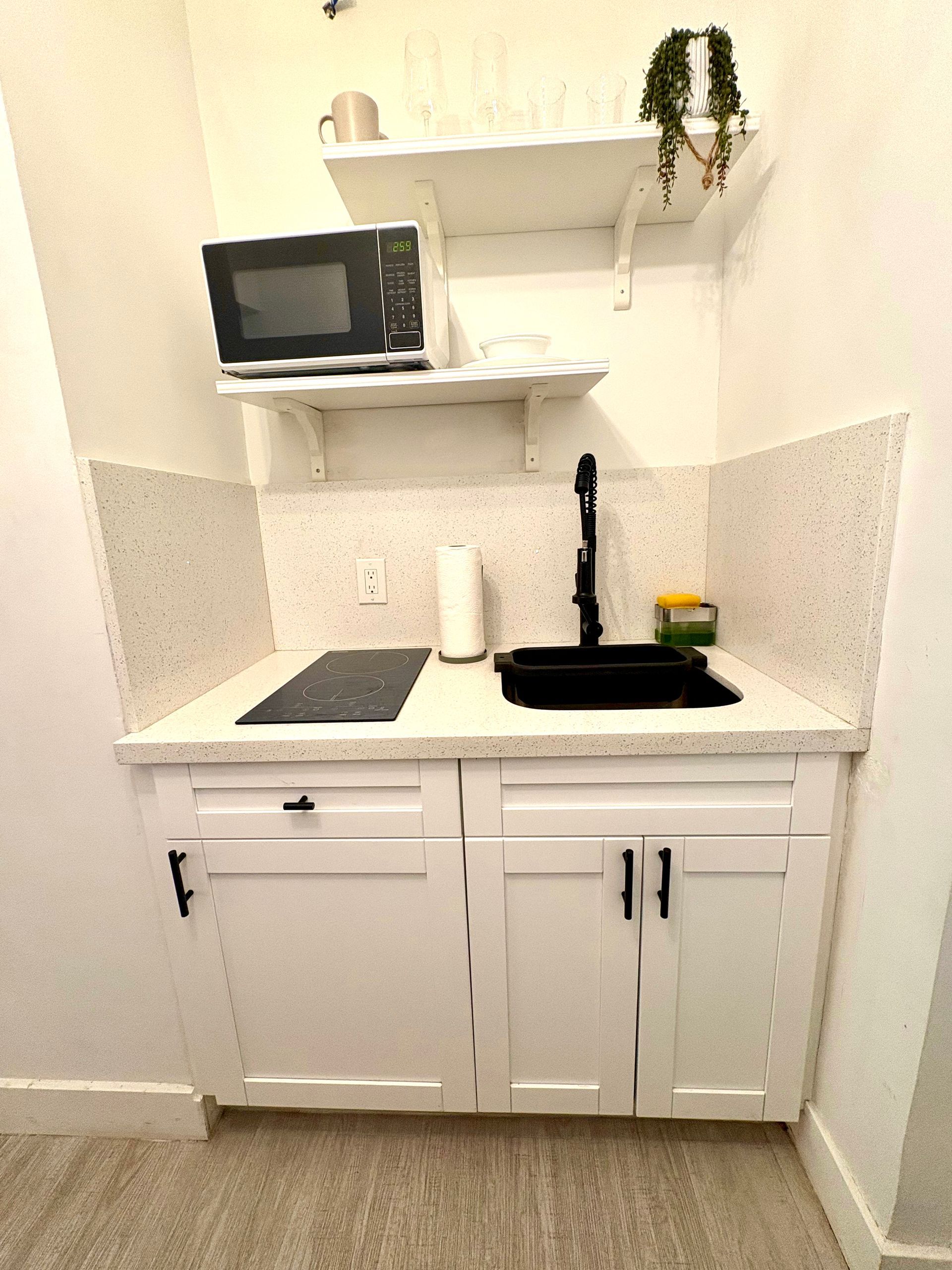 Small, white kitchenette with microwave, sink, and cabinets. A shelf holds glasses and a plant.