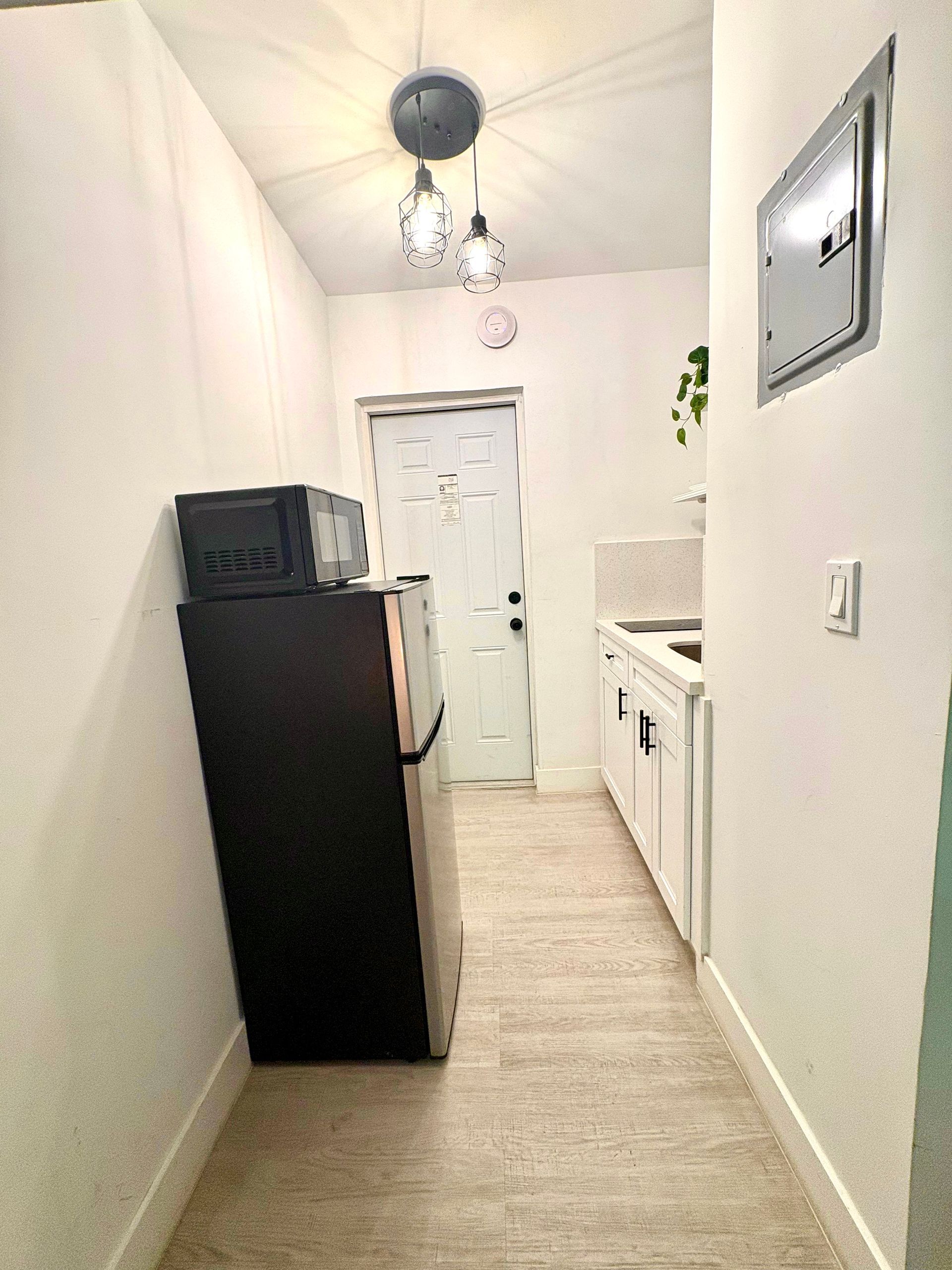 Narrow kitchen with refrigerator, microwave, sink, and door at end; white walls and cabinets, wood floor.