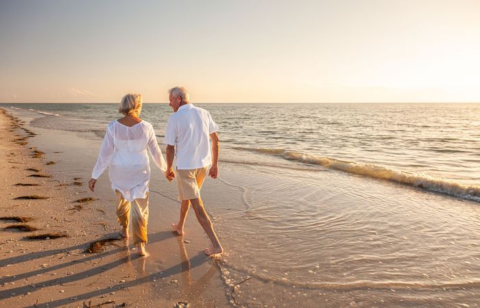 An elderly couple is walking on the beach holding hands.