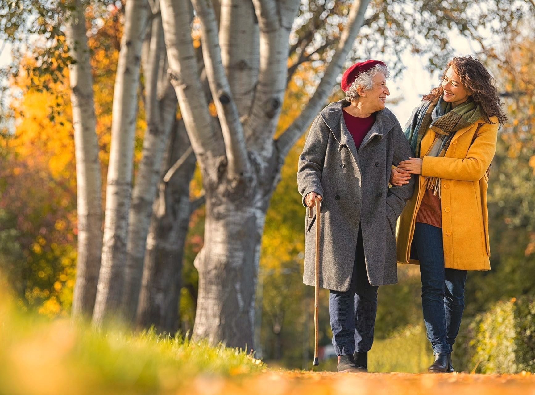 An older woman with a cane walks arm in arm with a younger woman along a tree-lined path in autumn sunlight.