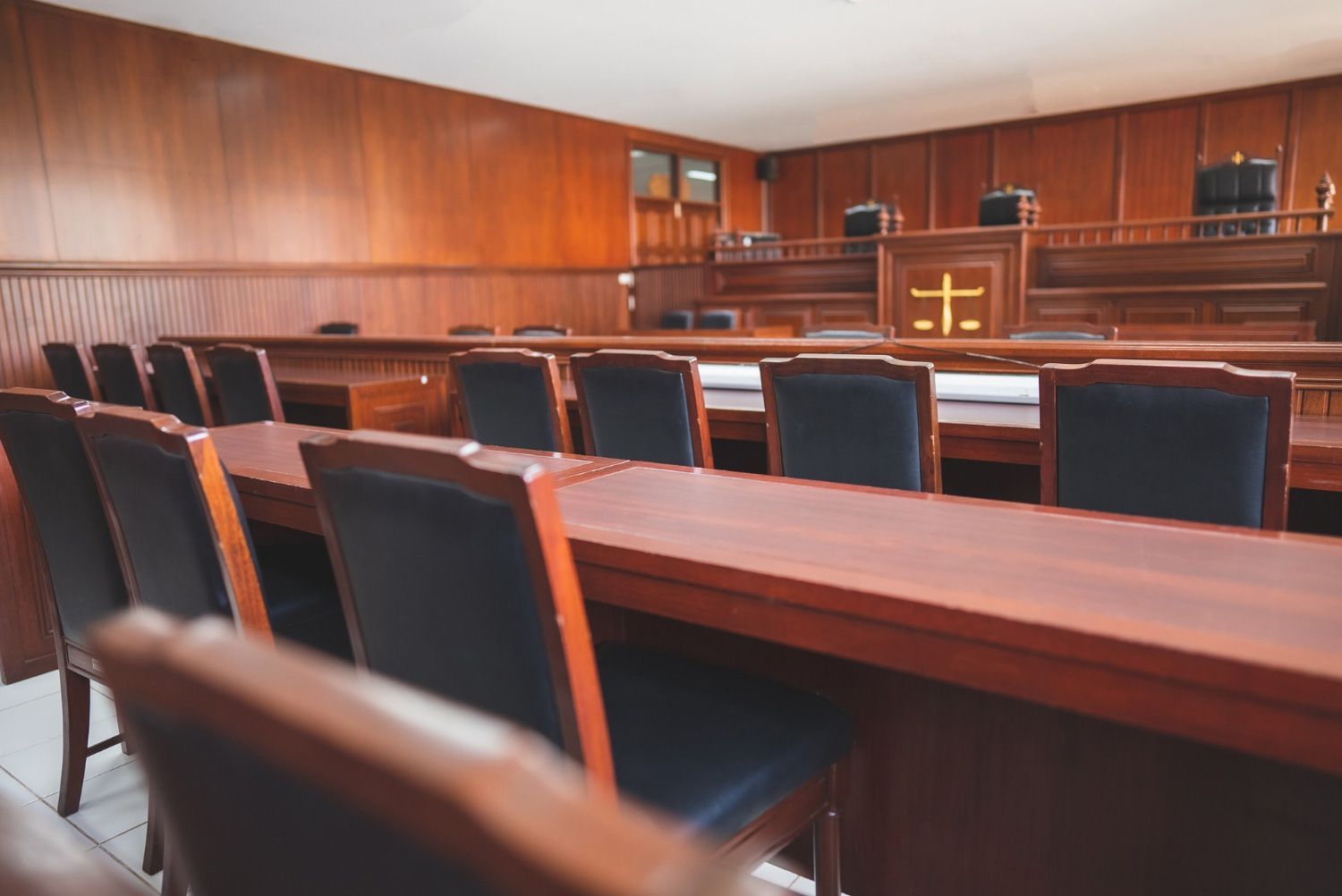 Empty wood-paneled courtroom with benches and chairs, viewed from the rear toward the judge’s bench.