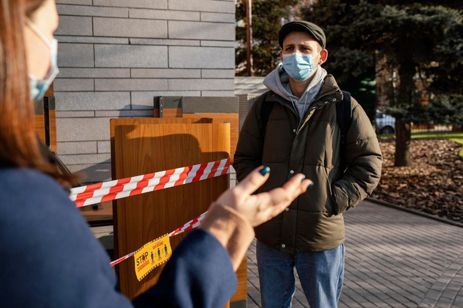Masked man speaks with someone behind red-and-white tape outside a building