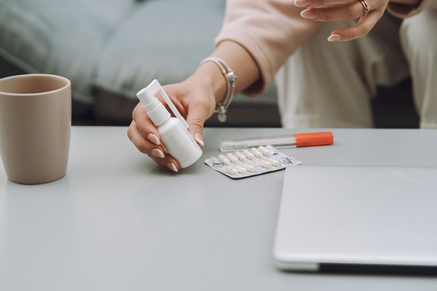 Hand pouring white pills from a bottle beside a laptop, mug, and blister pack on a table