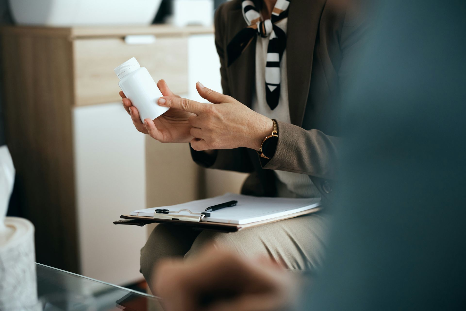 Person in a blazer holding a white pill bottle beside a clipboard in a meeting setting