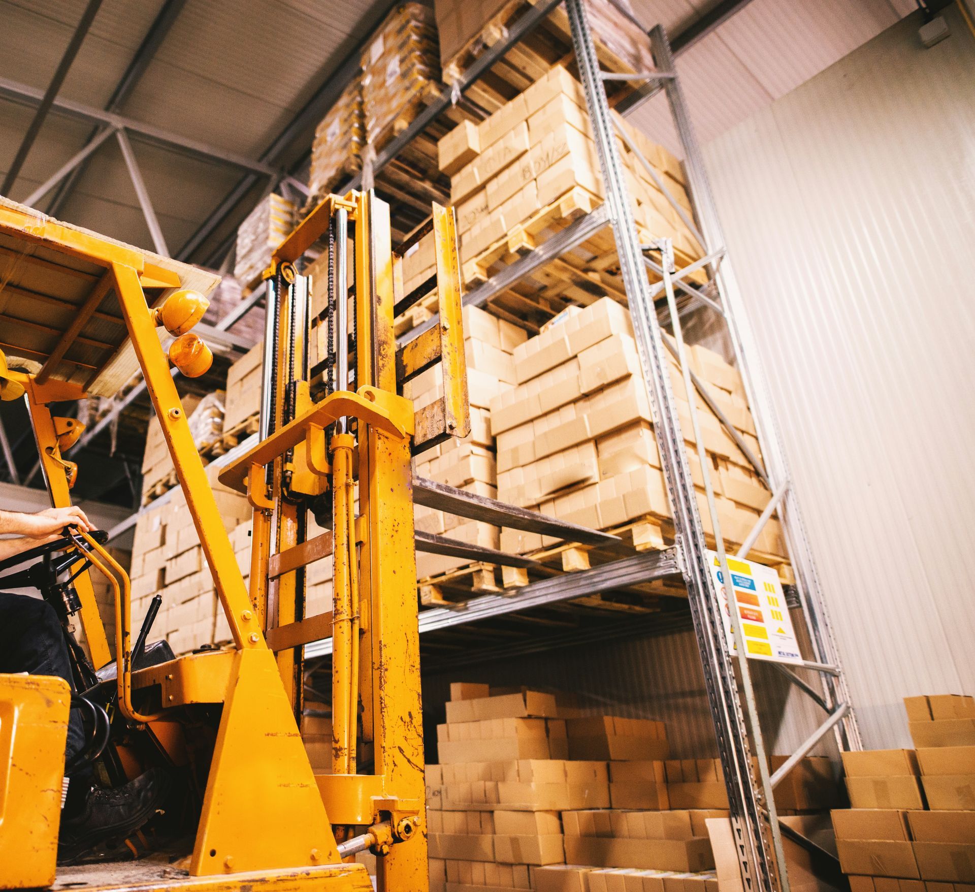 Forklift in a warehouse lifting stacked cardboard boxes on tall shelving, with more boxes stored below.