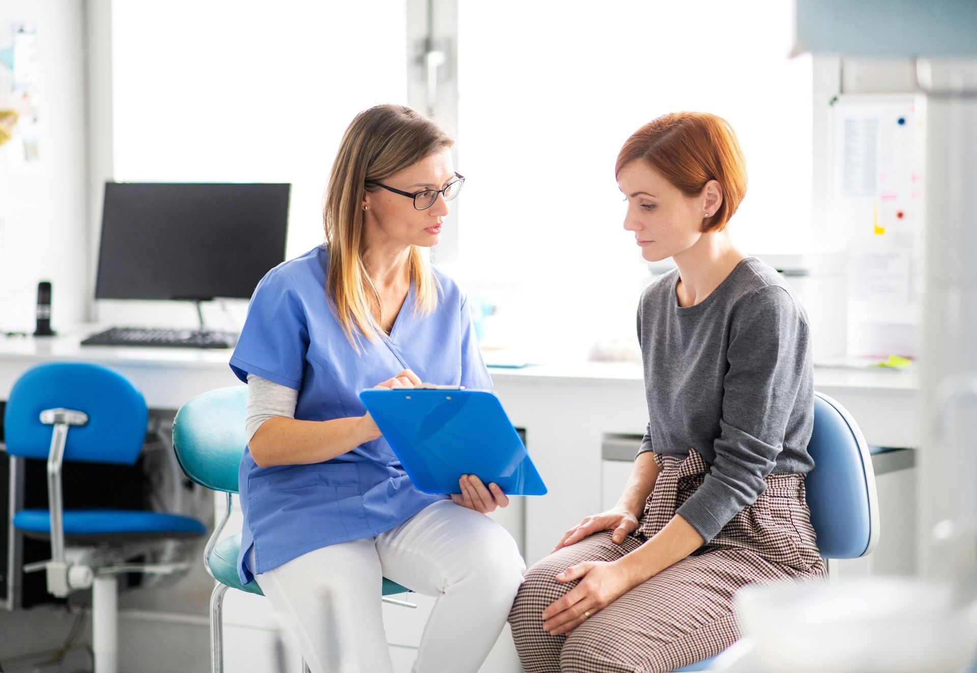 Two women talking in a bright office, one holding a blue clipboard.