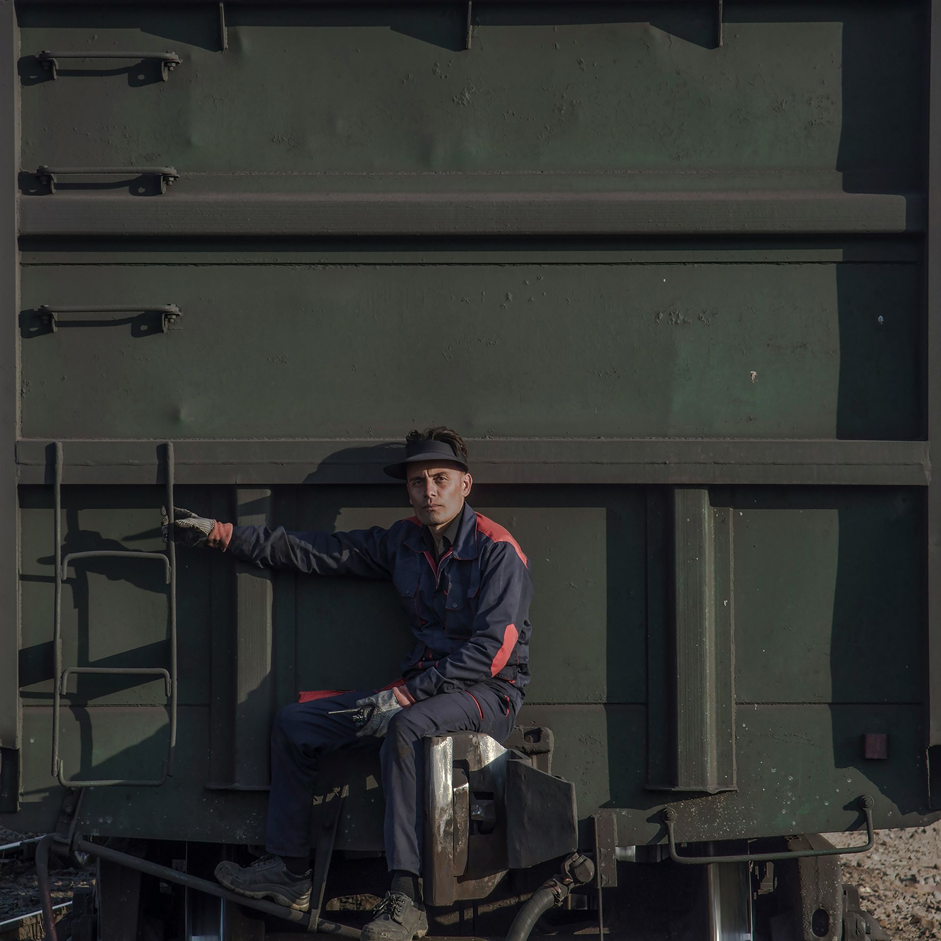Worker in dark uniform sitting on a green industrial machine, holding a ladder rail in a dim workshop