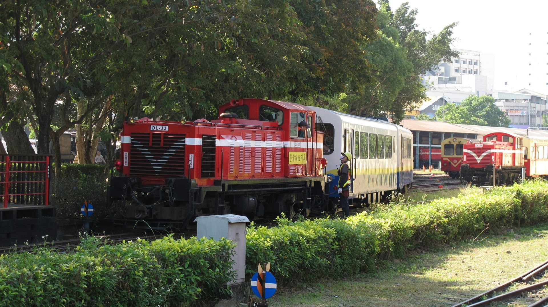 Red train engine pulling a yellow passenger train through a sunny, leafy park setting