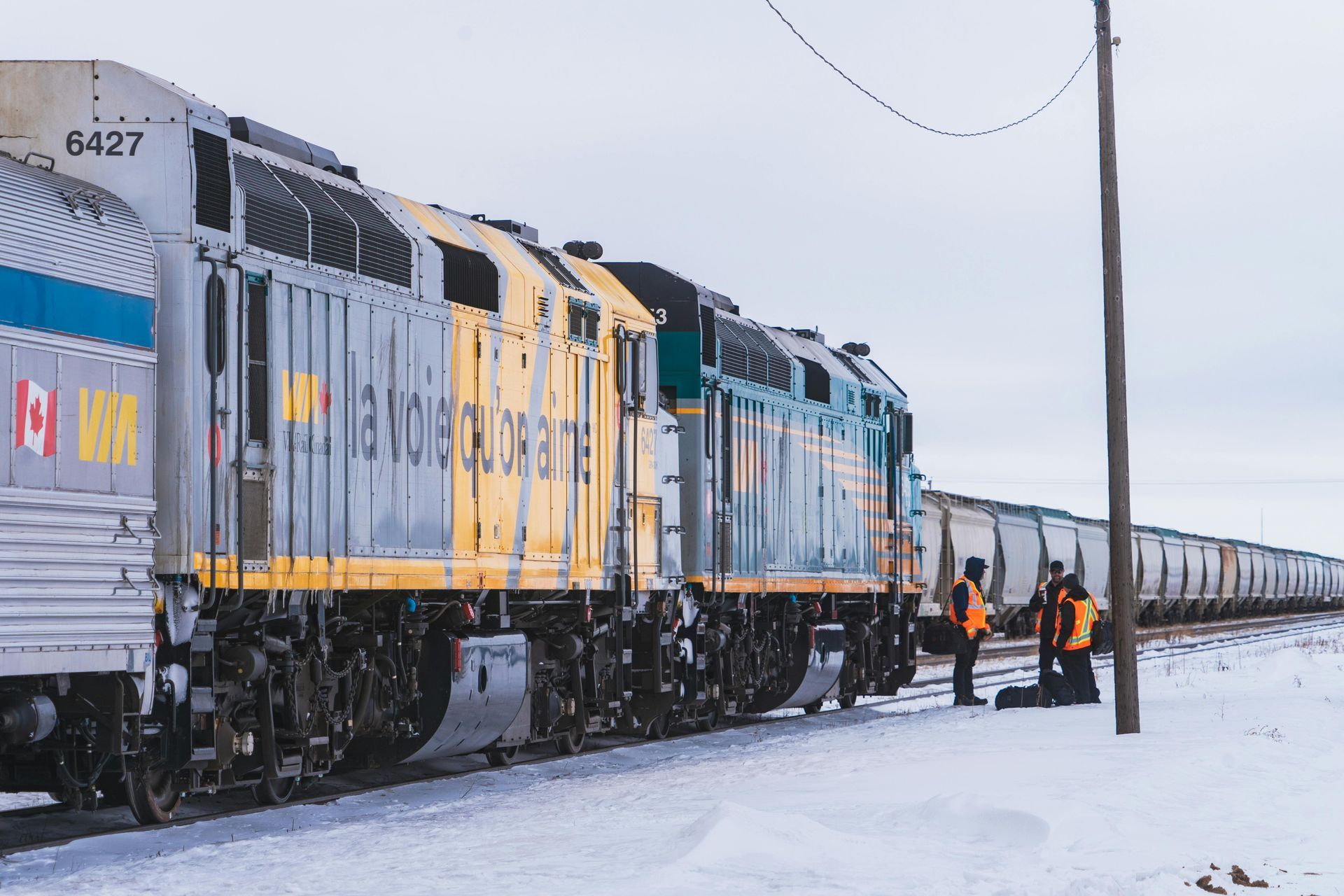 Freight locomotives on snowy tracks with workers beside a long train under a gray sky