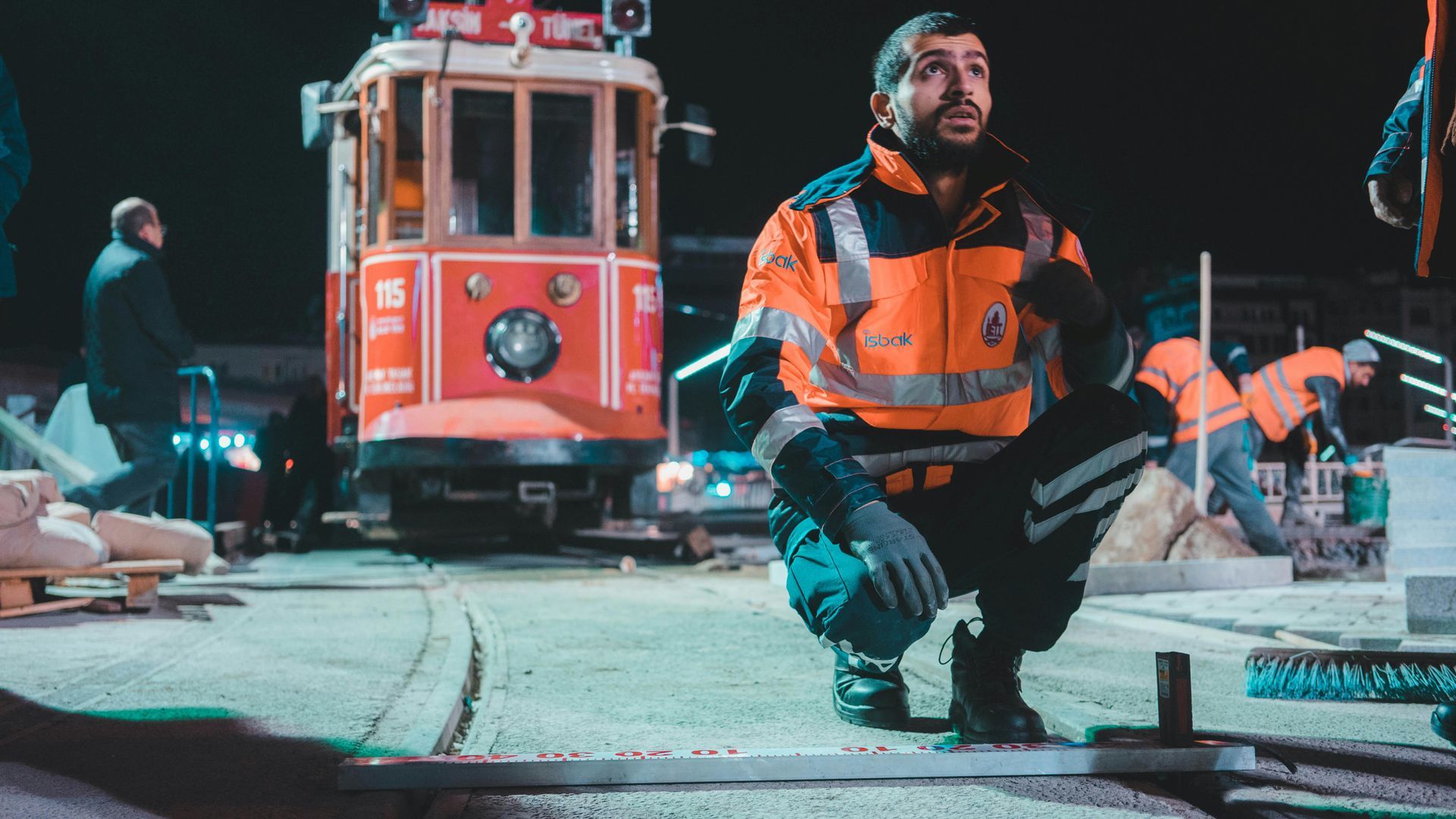 Worker in orange safety gear kneels on a street at night, with a red tram and equipment in the background.