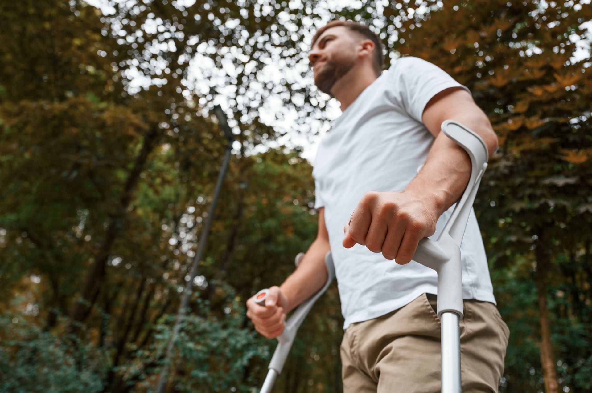 Man on crutches walking outdoors in a wooded area, wearing a white shirt and khaki pants.