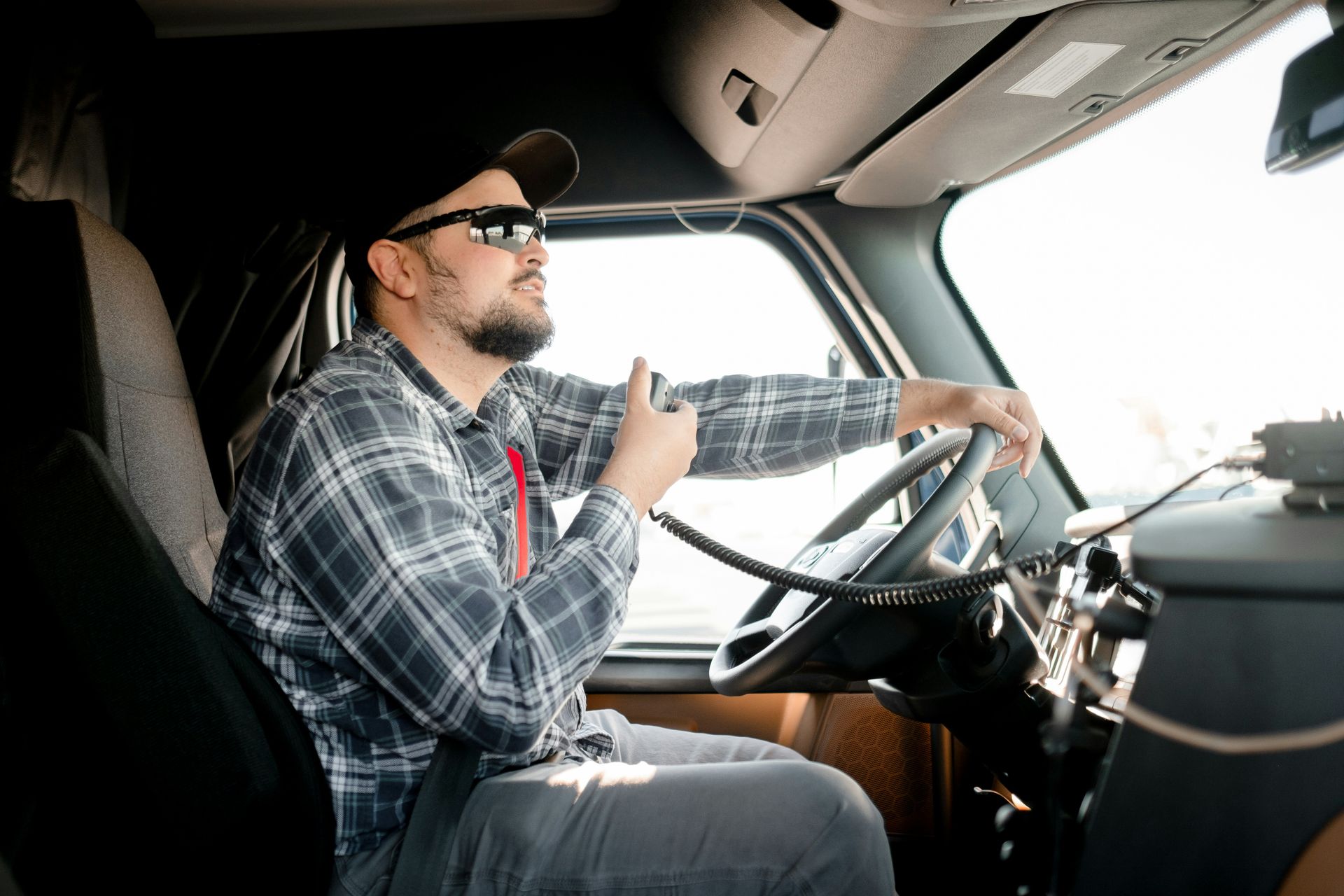 Driver in plaid shirt and cap steering a truck, with one hand on the wheel and sunlight through the window.