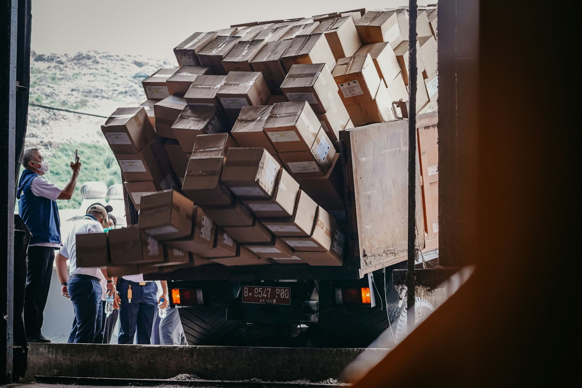 Workers guide a large wooden cube sculpture on a truck, viewed through a doorway.