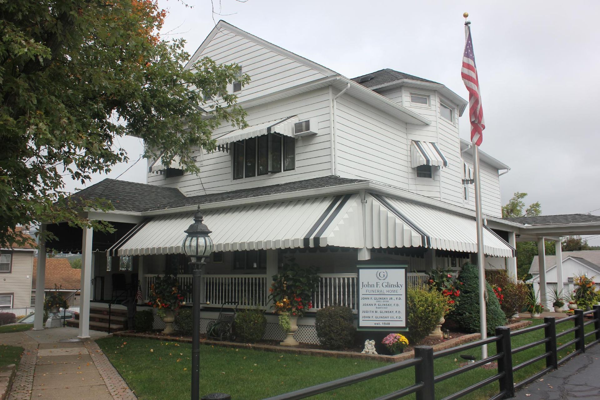 White house with black-and-white striped awning, American flag, and garden fence on a rainy street