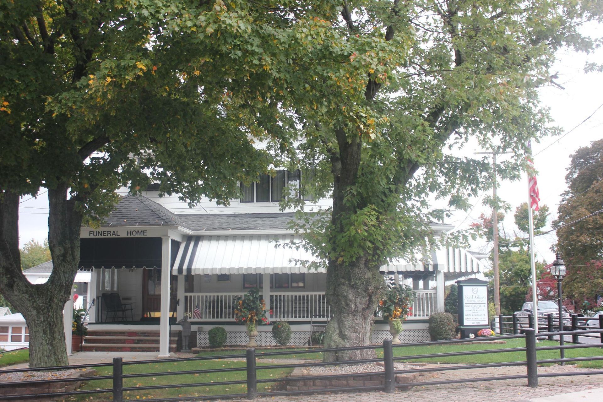 Tree-shaded street view of a white building with striped awnings and a fenced sidewalk