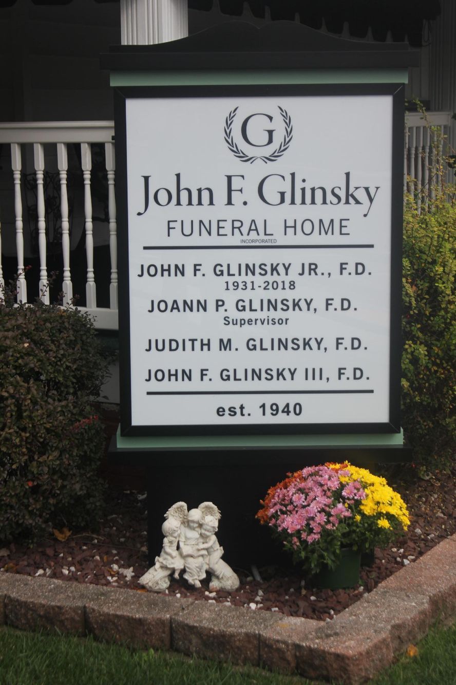 John F. Glinsky Funeral Home sign beside flowers and small memorial statue outdoors