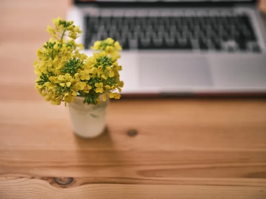 Small white vase of yellow flowers on a wooden table with a blurred laptop in the background