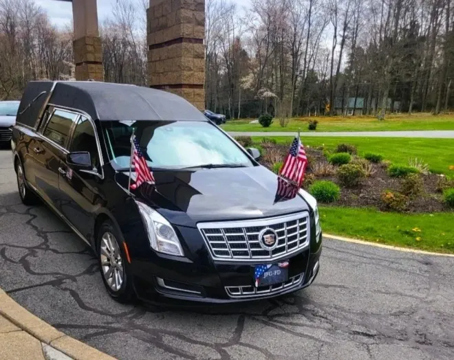 Black Cadillac hearse with U.S. flags parked on a road near a grassy park.