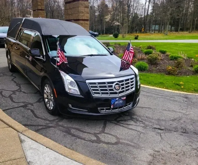 Black Cadillac hearse decorated with American flags parked on a wet driveway near a grassy cemetery.
