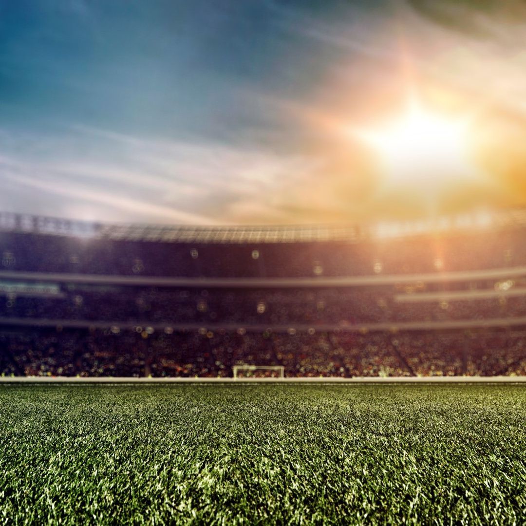 A green soccer field in a stadium, with a bright sun overhead and crowd blurred in the background.