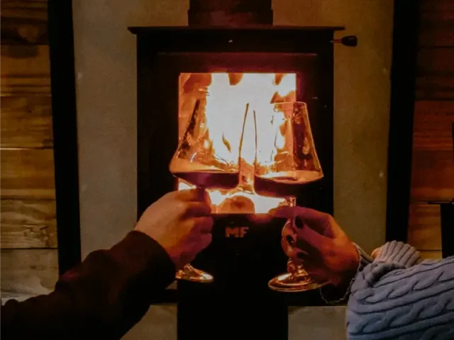 Duas mãos brindam com taças de vinho diante de uma lareira acesa, com uma lâmpada brilhante e uma sala aconchegante com painéis de madeira ao fundo.