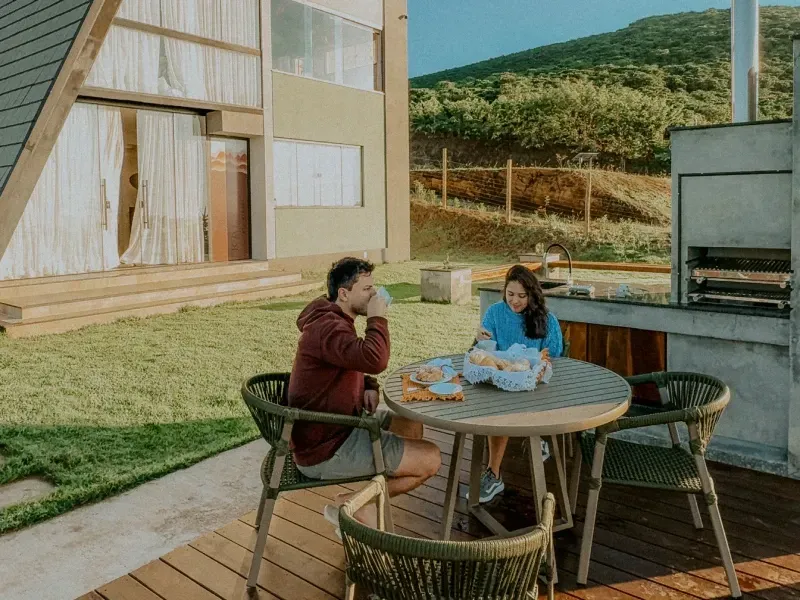 Duas pessoas conversando em uma mesa de madeira ao ar livre, ao lado de uma casa moderna e da vegetação da encosta.