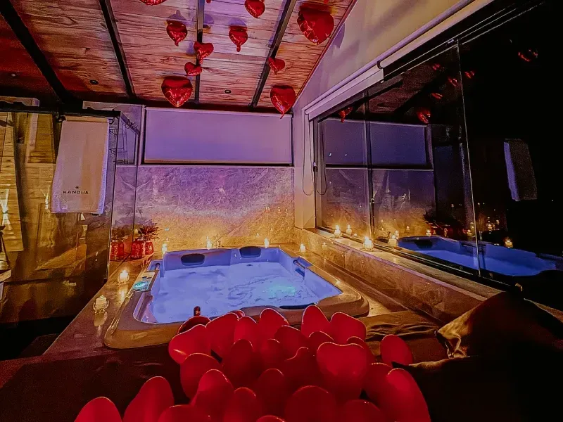 Banheira de hidromassagem Jacuzzi em uma sala com paredes de vidro, iluminada em roxo com balões vermelhos em primeiro plano.