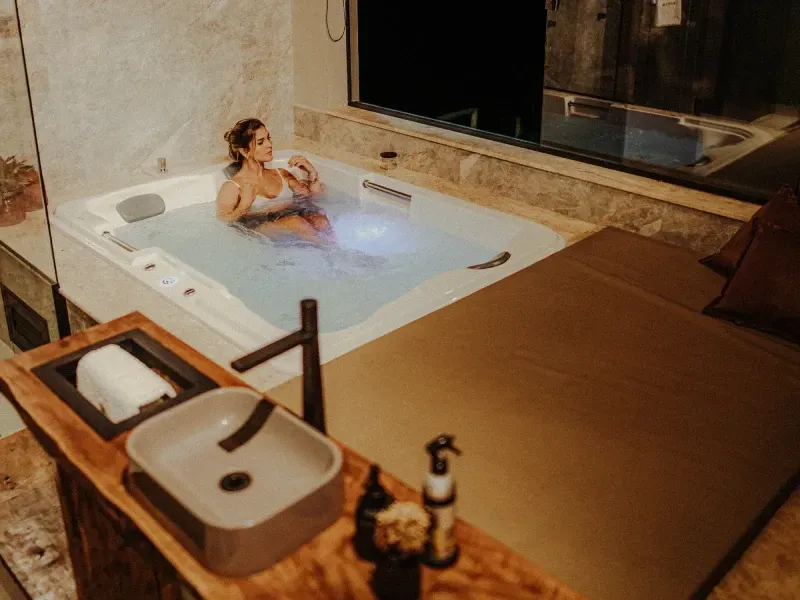 Pessoa relaxando em uma banheira de hidromassagem interna iluminada, ao lado de almofadas bege e uma parede de pedra.