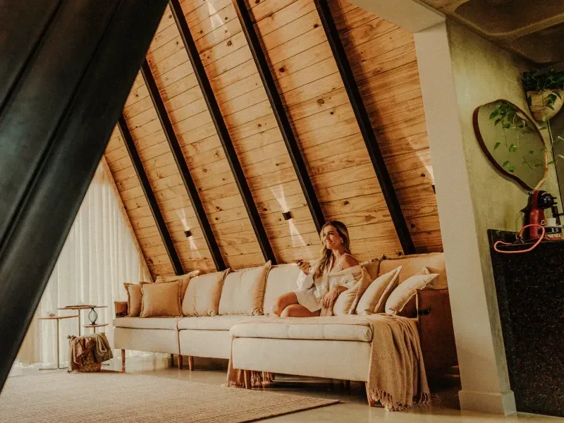 Sala de estar aconchegante em um loft com um sofá modular bege sob um teto inclinado de madeira e uma mulher sentada no sofá.