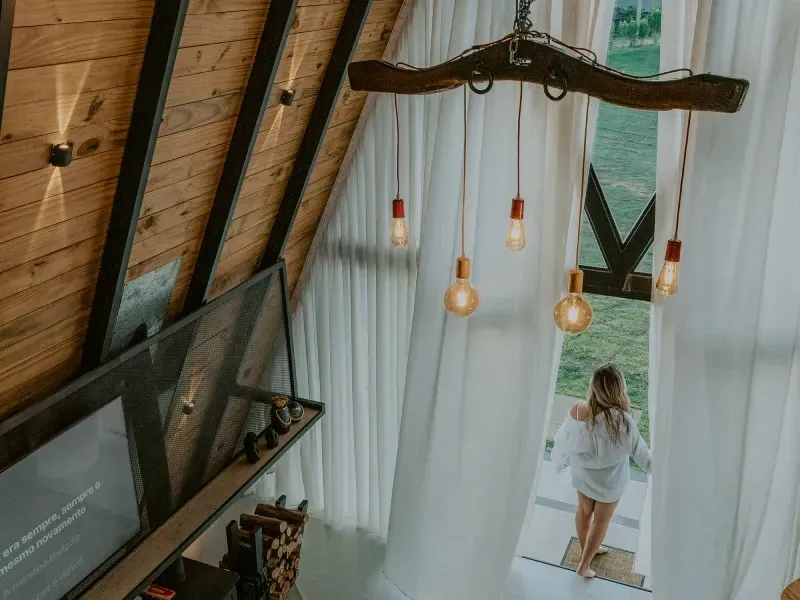 Mulher em pé num loft iluminado, perto de cortinas translúcidas, com luminárias pendentes e uma TV na parede.