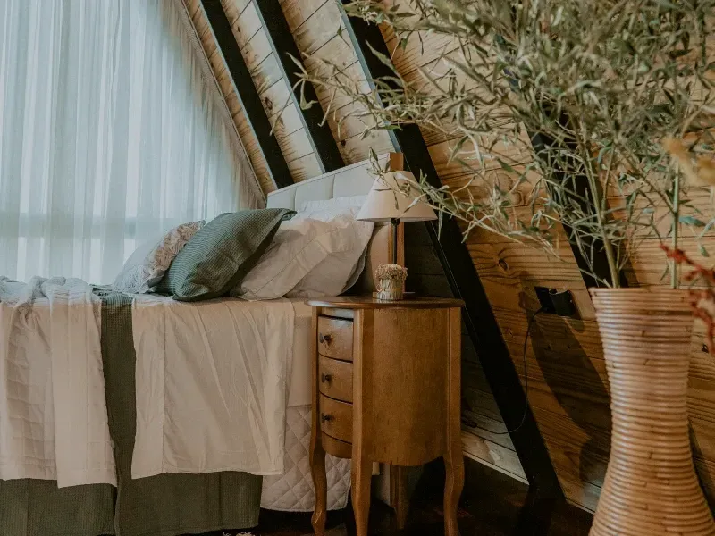 Quarto aconchegante com cama, mesa de cabeceira, janela grande e vaso alto de vime ao lado de uma planta frondosa.
