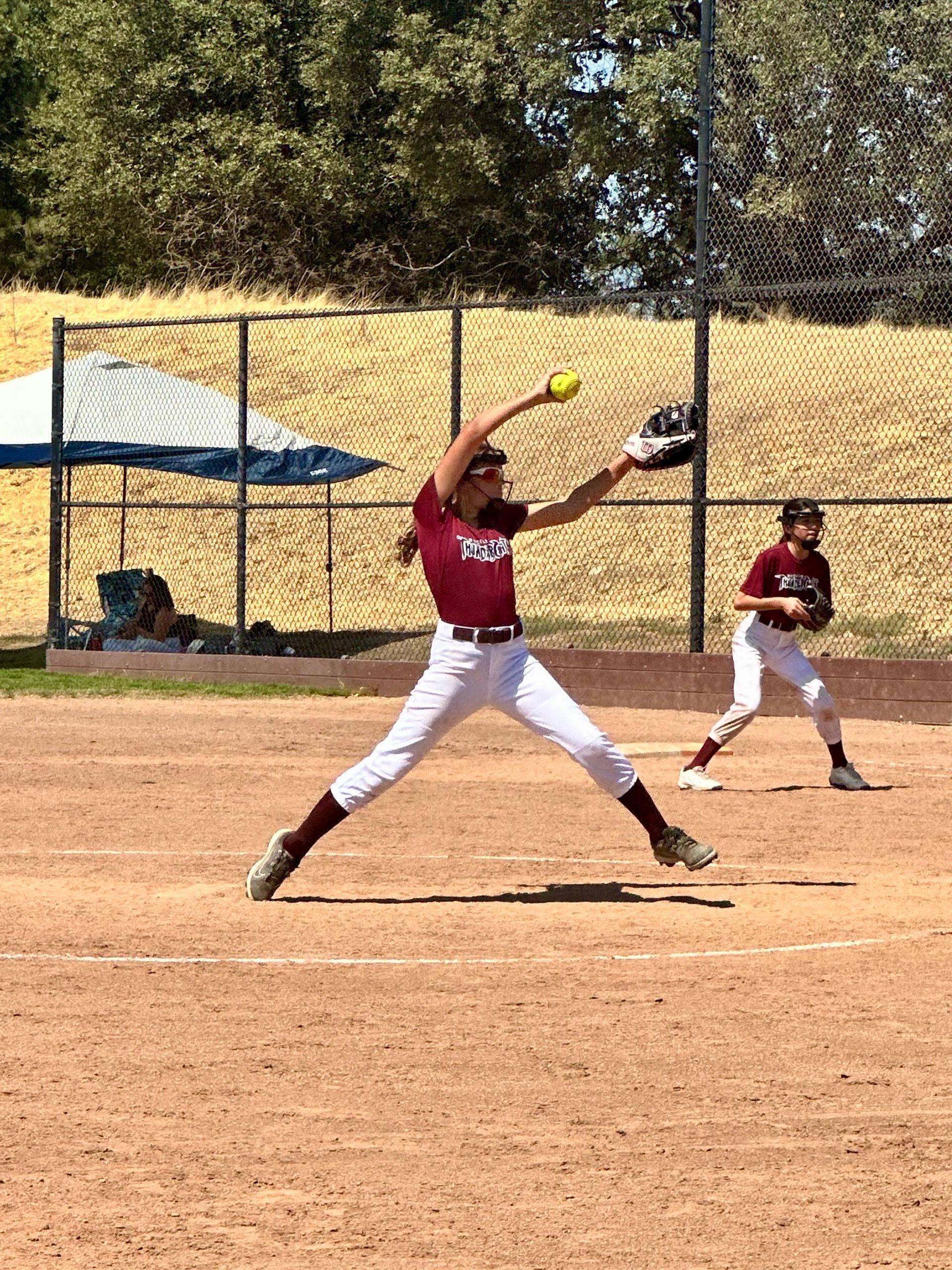 A softball pitcher in a maroon jersey and white pants winds up to throw a yellow ball on a dirt field.