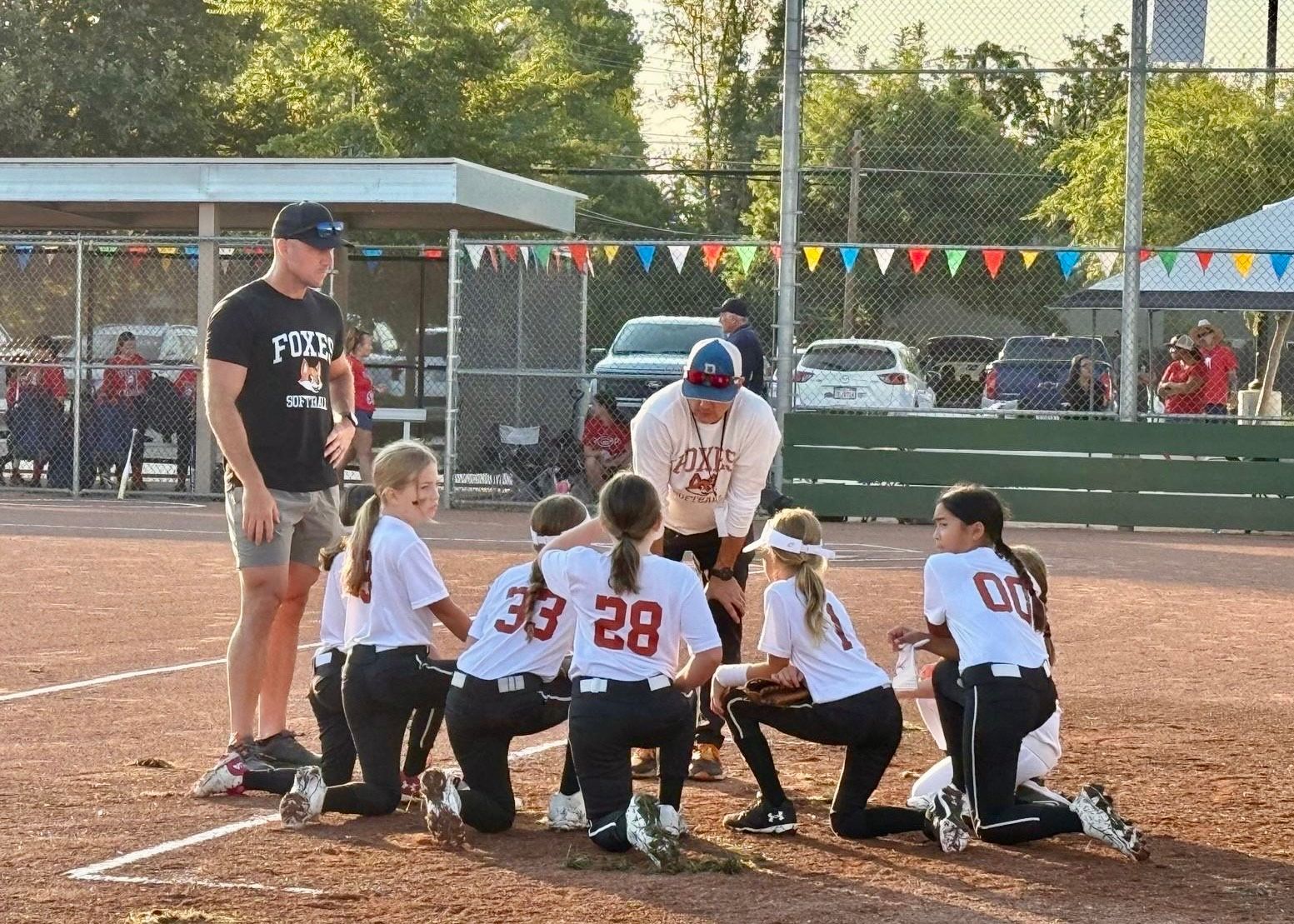 A coach and assistant standing over a circle of softball players kneeling in white uniforms on a dirt field.