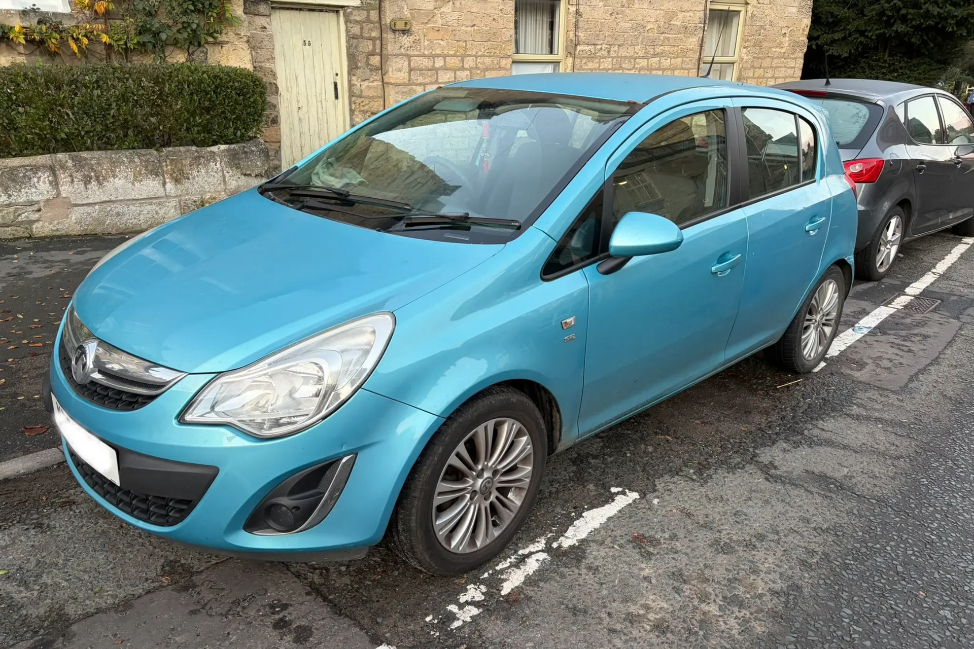 Light blue Vauxhall Corsa parked on a street. Grey car next to it, stone building in background.