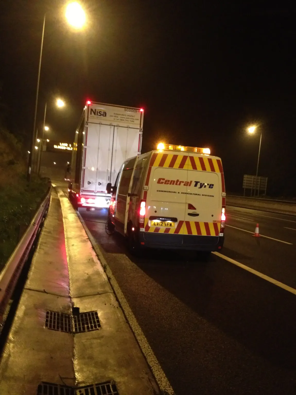 A Central Tyre service van with flashing lights next to a truck on the side of a road at night.