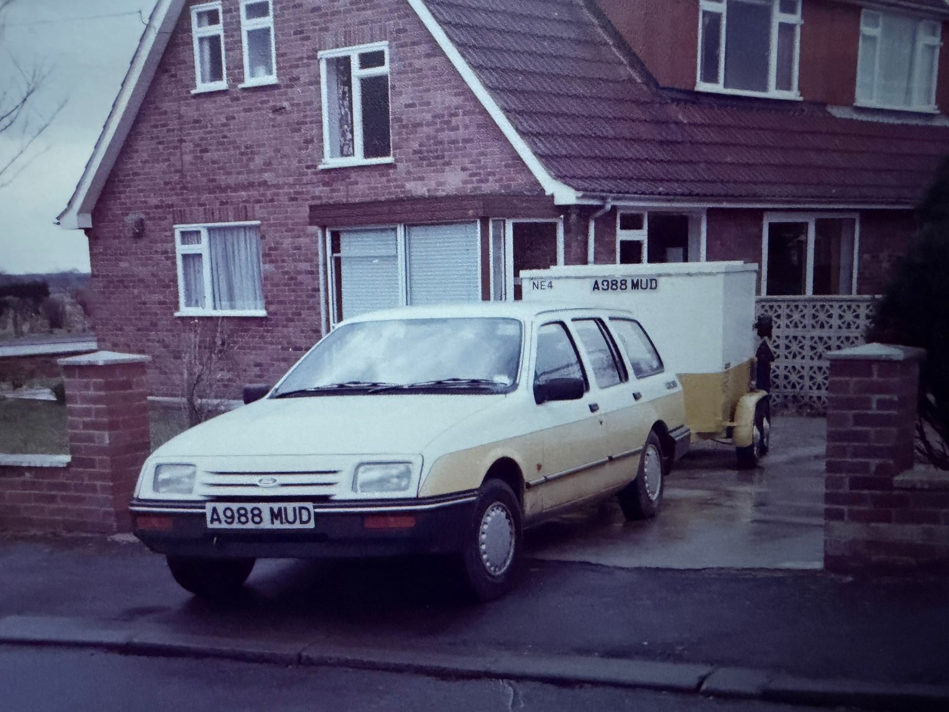 Cream-colored Ford Sierra estate car with a trailer parked in front of a brick house.
